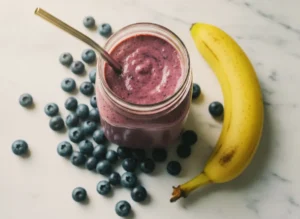 Banana blueberry smoothie in a glass mason jar with stainless steel straw, surrounded by fresh blueberries and banana on white marble