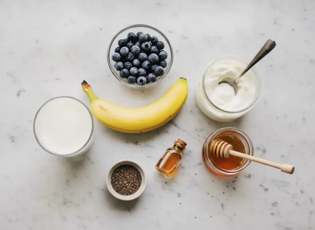 Banana Blueberry Smoothie Recipe – Thick, Creamy & Ready in 5 Minutes 2 Banana blueberry smoothie ingredients flat lay including frozen blueberries, banana, Greek yogurt, honey, and almond milk on white marble